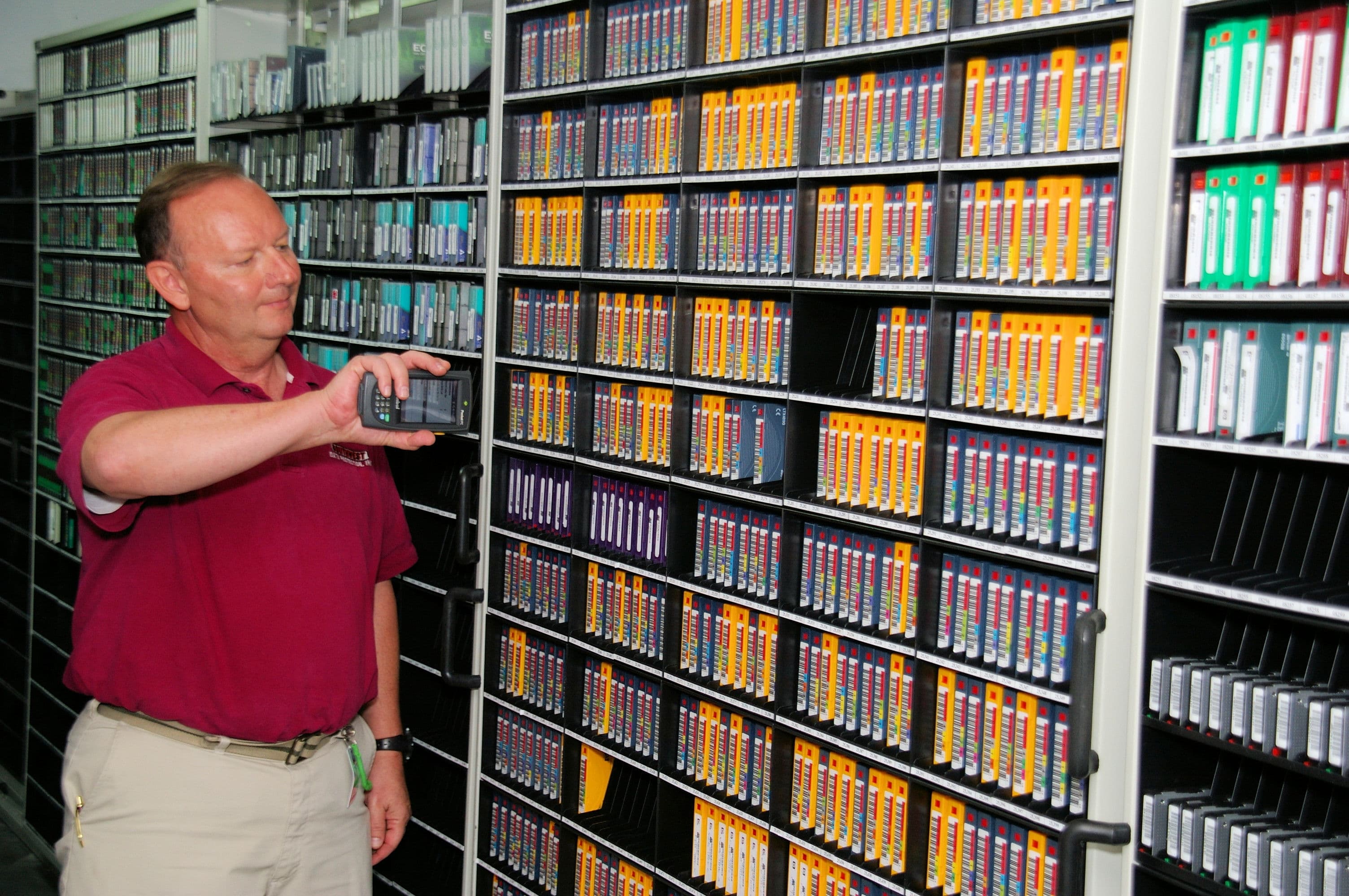 Man in a red shirt scanning labeled data tapes in a secure, organized tape vault storage facility.
