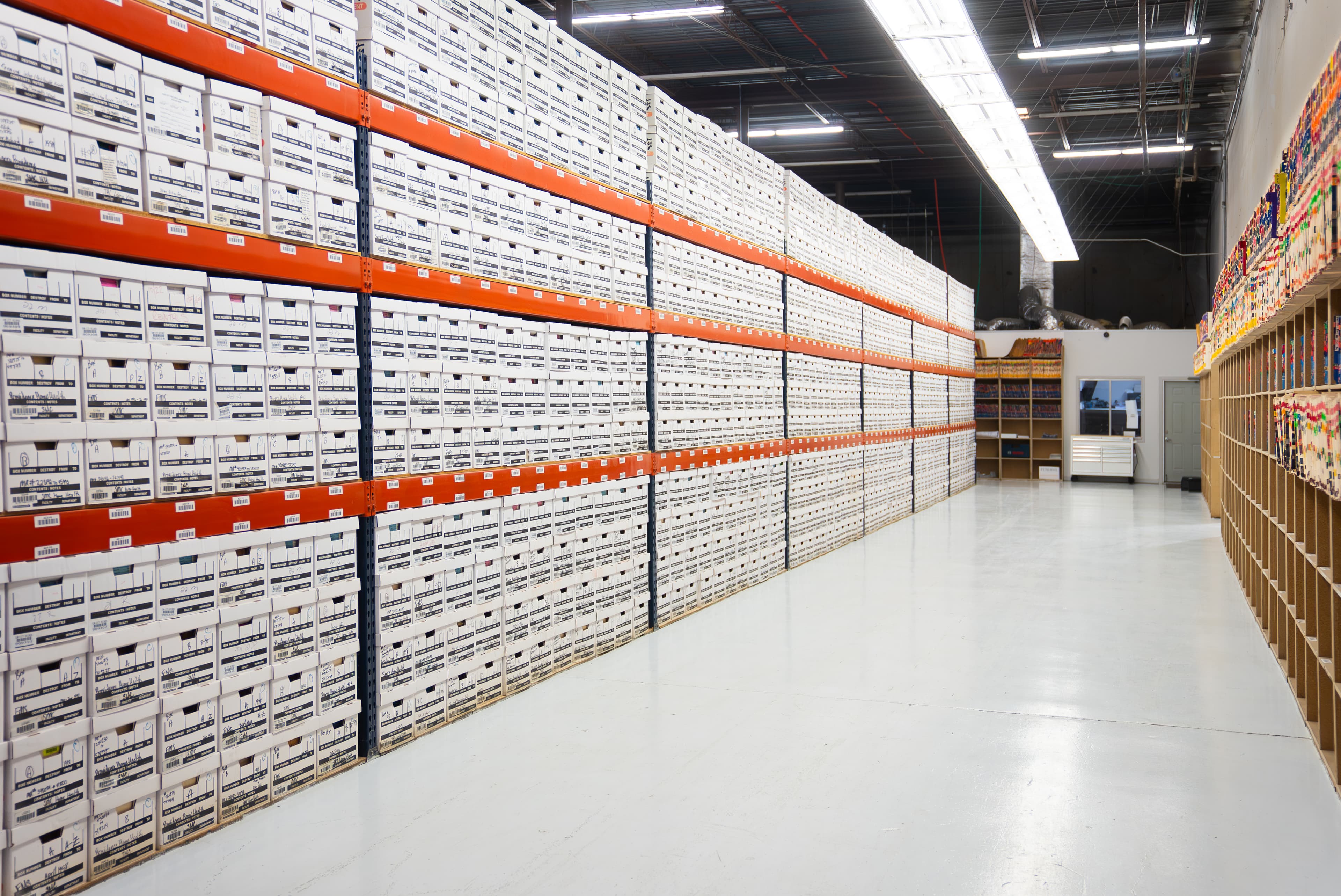 Interior view of a secure records storage facility, showing rows of stacked white archive boxes on industrial shelving with orange beams, under bright overhead lighting.